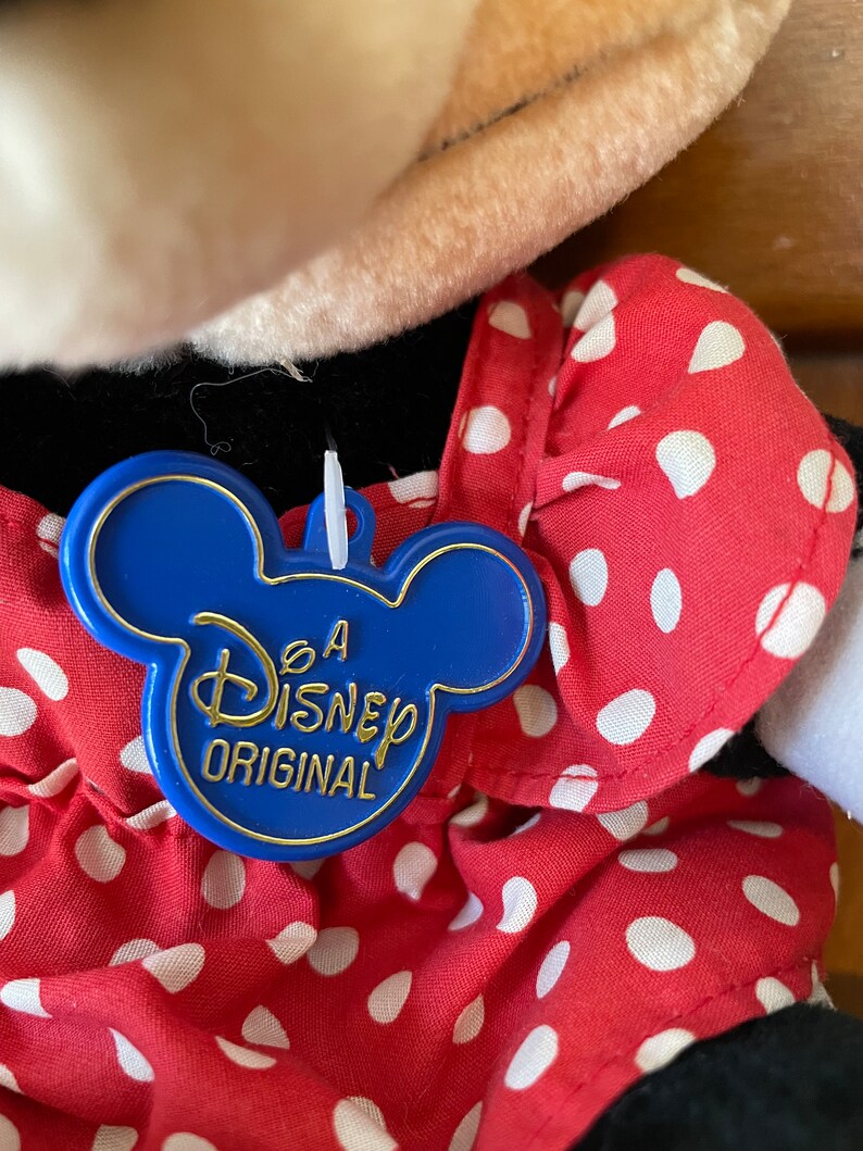 Pu&ograve; includere: Un'etichetta di plastica blu a forma di testa di Topolino con il testo "A Disney Original" in lettere dorate. L'etichetta &egrave; attaccata a un vestito rosso con pois bianchi.