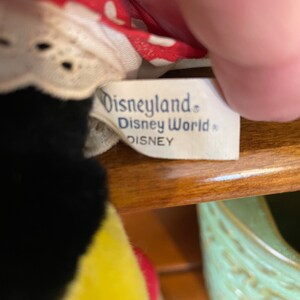 Pu&ograve; includere: Primo piano di un'etichetta bianca con testo blu che recita "Disneyland. Disney World. DISNEY". L'etichetta &egrave; attaccata a un tessuto a pois rosso e bianco con un bordo di pizzo bianco.