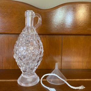 May include: A clear glass oil lamp with a textured, bulbous body and a handle. The lamp comes with a clear plastic funnel and a white wick.