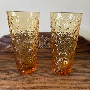 May include: Two tall drinking glasses made of amber-colored glass. Each glass features a distinct, heavy, textured surface with an irregular, organic pattern that resembles cracked ice or molten glass, creating a tactile and reflective appearance.