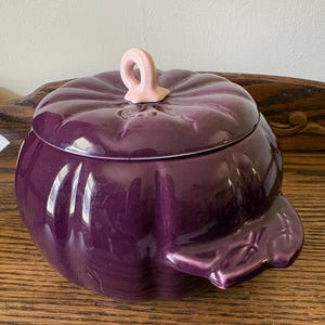 May include: A deep purple ceramic lidded bowl shaped like a pumpkin with a glossy finish. The lid features a light pink stem-shaped handle, and the bowl includes a small, sculpted leaf-shaped handle on the side, mimicking the natural form of a gourd.