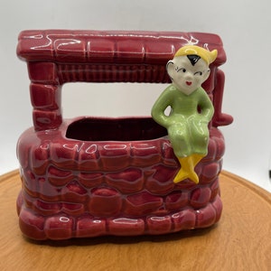 May include: A red brick well planter with a green and yellow elf figurine sitting on the edge. The elf is wearing a yellow hat and has a black face.