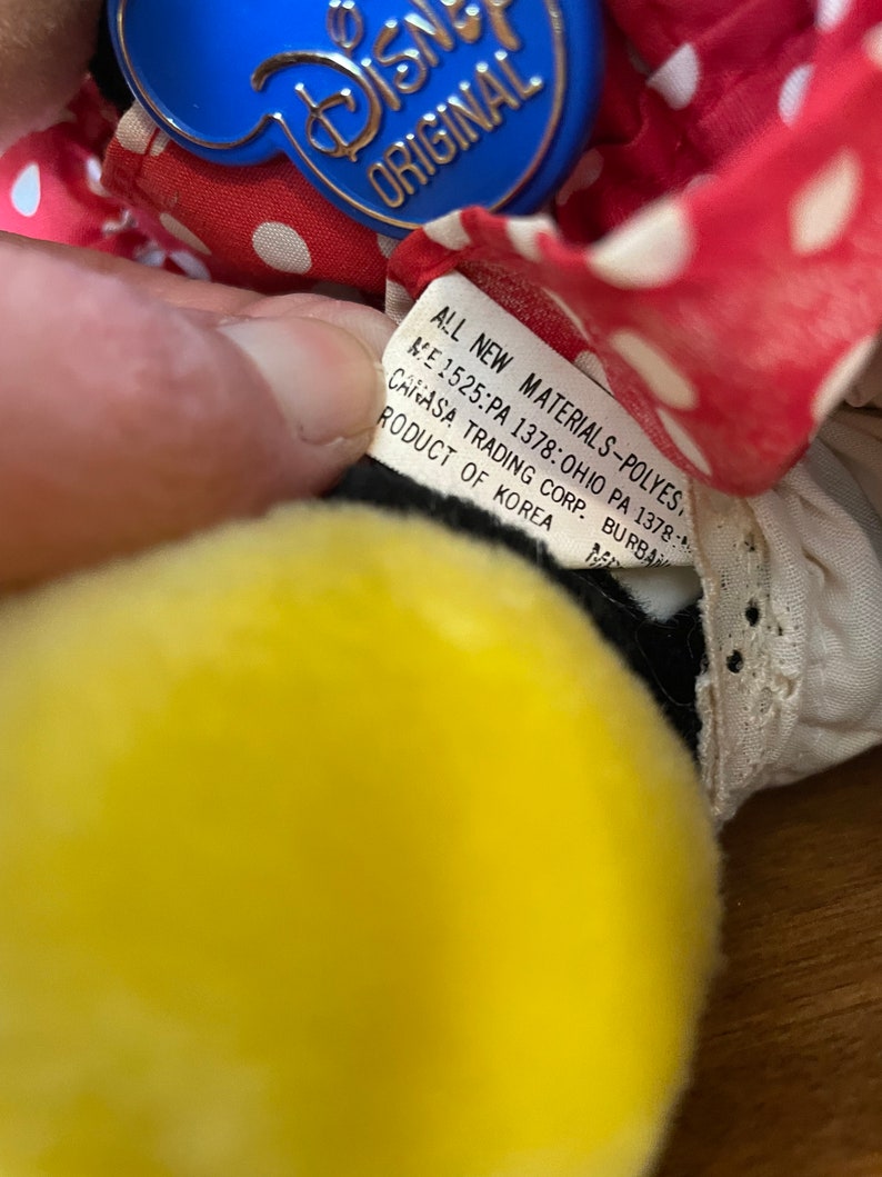 Pu&ograve; includere: Una bambola di stoffa a pois rossi e bianchi con una testa gialla e occhi neri. La bambola indossa un colletto di pizzo bianco e un'etichetta blu Disney Original attaccata al colletto. L'etichetta recita "ALL NEW MATERIALS-POLYES. ME 1525:PA 1378: OHIO PA 1378: CANASA TRADING CORP. BURBA RODUCT OF KOREA."