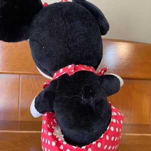 Pu&ograve; includere: Un giocattolo di peluche nero e rosso a forma di topo dei cartoni animati che indossa un vestito a pois rossi. Il giocattolo &egrave; seduto su uno scaffale di legno.