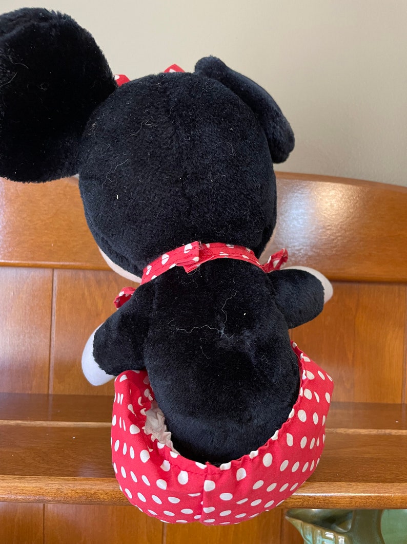 Pu&ograve; includere: Un giocattolo di peluche nero e rosso a forma di topo dei cartoni animati che indossa un vestito a pois rossi. Il giocattolo &egrave; seduto su uno scaffale di legno.
