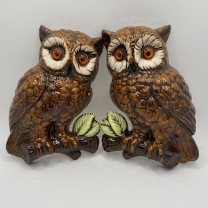May include: Two brown ceramic owls perched on branches with green leaves. The owls have white and orange eyes and brown and white feathers.