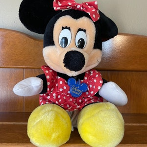 Pu&ograve; includere: Un giocattolo di peluche di Minnie Mouse che indossa un vestito rosso con pois bianchi e un fiocco rosso con pois bianchi. Il giocattolo ha un'etichetta blu che dice "A Disney Original".