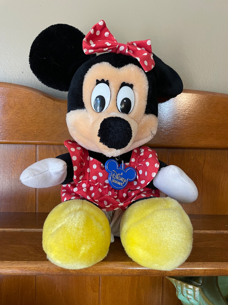 Pu&ograve; includere: Un giocattolo di peluche di Minnie Mouse che indossa un vestito rosso con pois bianchi e un fiocco rosso con pois bianchi. Il giocattolo ha un'etichetta blu che dice "A Disney Original".