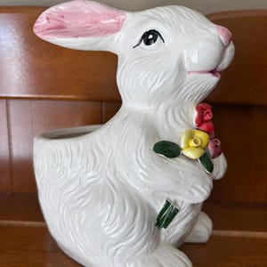 May include: A white ceramic bunny planter with pink accents on the ears and nose. The bunny holds a cluster of colorful flowers. The planter has a hollowed-out area for plants or decorations, perfect for Easter.