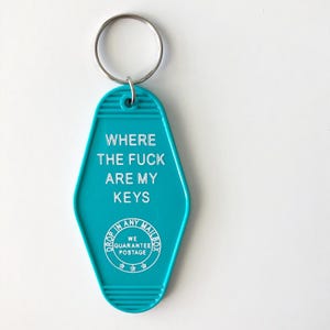 以下が含まれることがあります： 白い文字で「WHERE THE FUCK ARE MY KEYS」と書かれた、ターコイズ色のキーホルダー。キーホルダーにはシルバーのキーリングと、「DROP IN ANY MAILBOX WE GUARANTEE POSTAGE」と書かれた小さな円形のロゴが付いています。