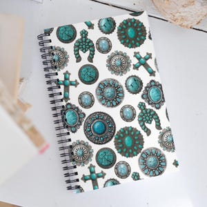 May include: Spiral-bound notebook with a white cover featuring a repeating pattern of turquoise and silver-toned decorative elements. The design includes crosses, ovals, and circular shapes, creating a Southwestern or bohemian aesthetic.