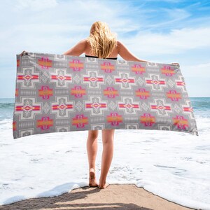 May include: A gray beach towel with a pink, orange, and white geometric pattern. The towel is being held by a person standing on a sandy beach.