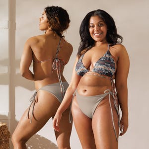 May include: Two women wearing a patterned bikini top and plain coloured bikini bottoms. The bikini top has a geometric pattern in shades of blue, red, and white. The bikini bottoms are a solid light grey colour with ties on the sides.