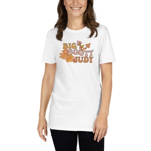 Op de afbeelding: Wit T-shirt met de tekst "BIG BOOTY JUDY" in oranje en gele retro letters, vergezeld van bloemen- en paddenstoelafbeeldingen. Het shirt heeft een ronde hals en korte mouwen.