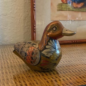 May include: A decorative duck ornament with a metallic finish. The duck has a brown head and beak, and its body is decorated with detailed floral patterns in silver, orange, and green. The ornament is placed on a woven surface.