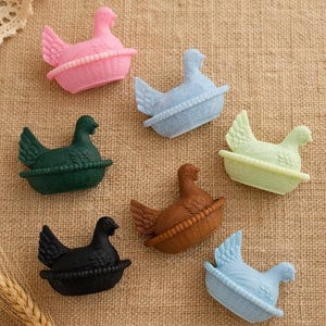 May include: A selection of colourful hen figurines, each in a basket-like base. The hens are in various colours, including pink, blue, green, brown, and black. The image is set against a textured, neutral background.