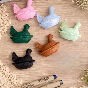 May include: A collection of colorful hen figurines in pink, blue, green, orange, and black, each nestled in a basket. The figurines are arranged on a light wood surface, with two pens and decorative elements.