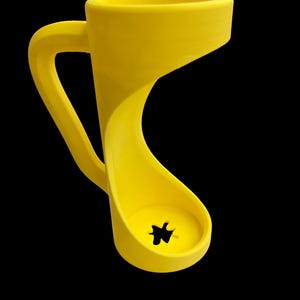 May include: A bright yellow, uniquely designed mug with a curved handle and a distinctive, open-bottomed base. The mug's form is a continuous, flowing shape, with a star-shaped hole at the base. The background is solid black.