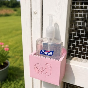 May include: A light pink square holder with a scalloped top edge and a rooster silhouette embossed on the front. The holder contains a clear plastic pump bottle of Purell hand sanitizer and is mounted to a white wooden structure with wire mesh.