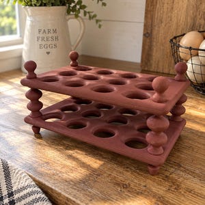 May include: A terracotta-colored egg holder featuring two stacked rectangular tiers with circular cutouts for eggs. The structure is supported by four decorative, spindle-shaped corner posts with rounded finials at the top and small feet at the base.