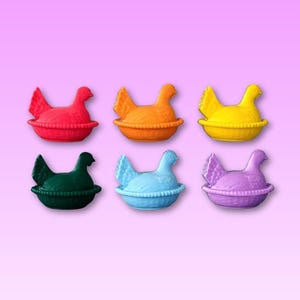May include: Six brightly coloured hen-shaped containers in red, orange, yellow, green, blue, and purple. Each hen sits in a basket-like base. The background is a pink to purple gradient.