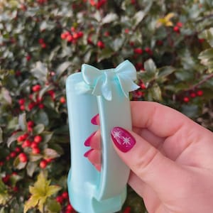 May include: A pale blue gift bag with a matching bow. The bag is held against a backdrop of green foliage and red berries. The bag has a handle and a bow at the top.