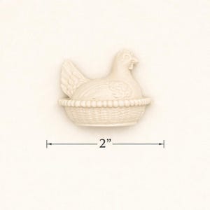 May include: A cream-coloured ceramic hen figurine, 5 cm in length, sitting in a woven basket. The hen has detailed feathers and a beak. The basket has a textured, woven design. The figurine is a decorative item.