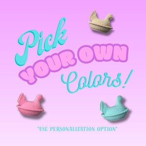 May include: Image shows three hen figurines in pink, blue, and beige, set against a gradient pink and purple background. Text reads "Pick YOUR OWN Colors!" with a note to use the personalization option.
