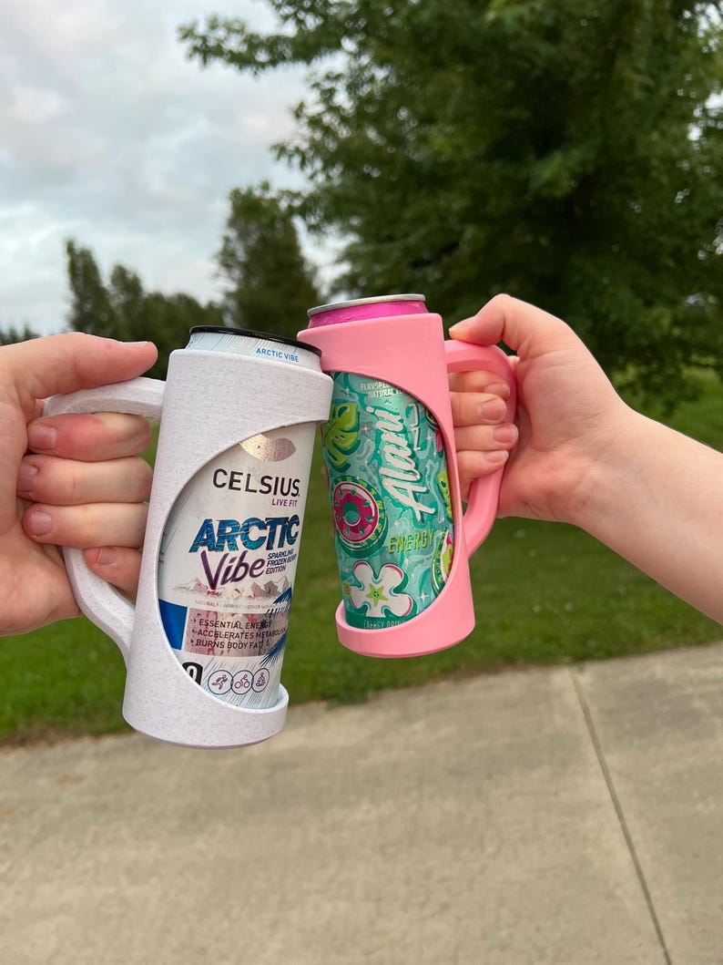 May include: Two beverage can holders, one white and one pink, each holding a can. The white holder has the text "CELSIUS ARCTIC VIBE" on the can. The pink holder has a can with a floral design and the text "Alani". Both holders have handles.