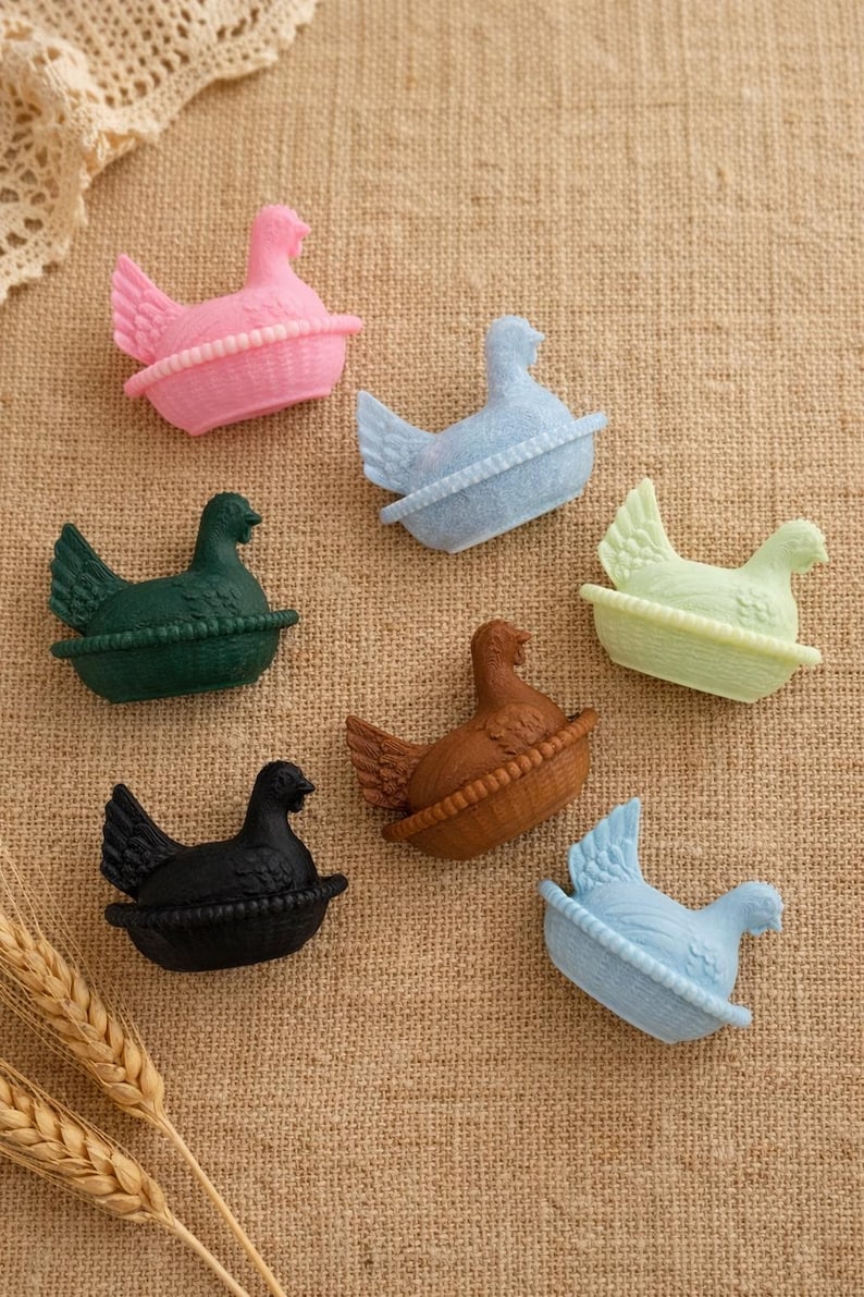 May include: A selection of colourful hen figurines, each in a basket-like base. The hens are in various colours, including pink, blue, green, brown, and black. The image is set against a textured, neutral background.