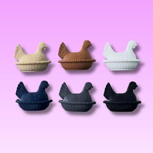 May include: Six ceramic hen ornaments in cream, brown, white, navy blue, grey, and black. Each hen sits in a woven basket. The background is a gradient of pink and purple.