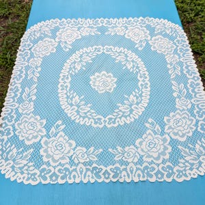 Op de afbeelding: Een vierkante, witte kanten doily met een bloemenontwerp. De doily heeft een centraal bloemmotief omgeven door een cirkelvormig patroon van bladeren en bloemen. De randen zijn geschulpt en versierd met meer bloemendetails. De doily ligt op een lichtblauwe ondergrond.