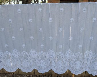 May include: A sheer, light gray curtain with a delicate white floral pattern. The curtain features a scalloped, ornate lace border along the bottom edge. The floral design is repeated throughout the fabric, creating a vintage or romantic aesthetic.