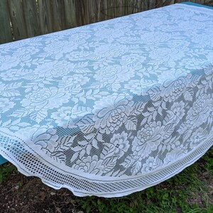 May include: White lace tablecloth with a floral pattern, draped over a blue table. The tablecloth has a scalloped edge and intricate floral designs. The table is outdoors, with a wooden fence and grass in the background.