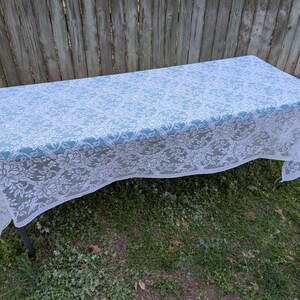 May include: A rectangular tablecloth with a white floral lace overlaying a light blue fabric. The tablecloth is draped over a table with dark metal legs, set on a grassy surface with a wooden fence in the background.