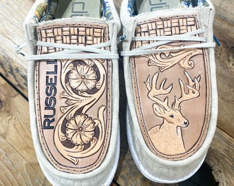 May include: Pair of beige slip-on shoes with leather accents. One shoe features a floral design and the name "RUSSELL", the other a deer. The interior lining has a floral pattern.