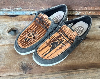 May include: A pair of gray canvas slip-on shoes with brown leather accents. The shoes feature a woven design on the leather and a graphic of a bear in a forest on one shoe and a graphic of a fist with a lightning bolt on the other shoe. The shoes have a white sole and the text "Hey Dude" is visible on the inside of the shoe.