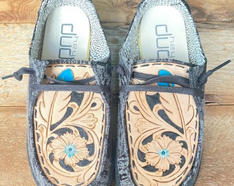 May include: Pair of gray slip-on shoes with a floral leather design. The shoes have a light tan leather floral pattern with a turquoise center. The insoles have the text "hey dude".