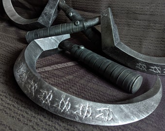 Op de afbeelding: Drie zilverkleurige metalen sikkel met zwarte handvatten. De sikkel zijn gebogen en hebben een scherpe rand. De handvatten zijn omwikkeld met zwart leer. De sikkel liggen op een bruine stoffen ondergrond op elkaar. De sikkel hebben markeringen op het blad die lijken op Japanse karakters.