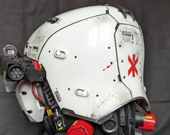Op de afbeelding: Witte helm met rode accenten en een rood kruis aan de zijkant. De helm heeft een zwart vizier en een zwarte band. De helm staat op een zwarte standaard.