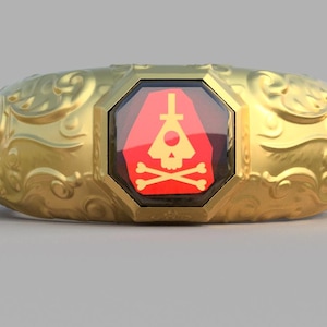 Könnte beinhalten: Ein goldener Ring mit einem verzierten Design und einem roten Edelstein, der in der Mitte eingesetzt ist. Der Edelstein zeigt einen Totenkopf und gekreuzte Knochen mit einem Dreieck und einem Kreis darüber.