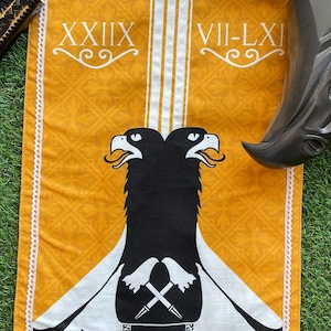 Op de afbeelding: Een witte banner met een zwart ontwerp van een dubbelkoppige adelaar. De banner heeft een gele achtergrond met een witte rand en een witte kantversiering. De adelaar heeft een zwart schild met een wit symbool erop. De banner heeft ook de Romeinse cijfers XXIII en VII-LXI.