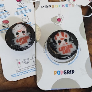 May include: Two PopSockets with a horror movie theme, featuring a skull design with red accents and the text "Crystal Lake." The PopSockets are on a white backing with the text "Friday the 13th."