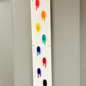 Puede incluir: Un lienzo rectangular blanco con nueve paletas de helado coloridas que gotean por la superficie. Las paletas de helado son rojas, naranjas, amarillas, verdes, azules, rosas, moradas y negras.