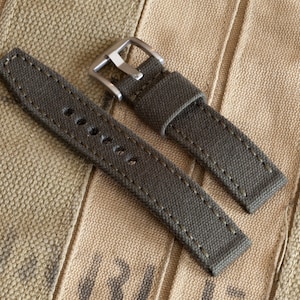 Puede incluir: Dos correas de reloj de lona verde oliva con cierres de hebilla plateados. Las correas están cosidas con hilo blanco.