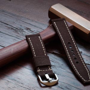 May include: Two brown canvas watch straps with white stitching. One strap has a buckle and the other has a series of holes for adjusting the fit.