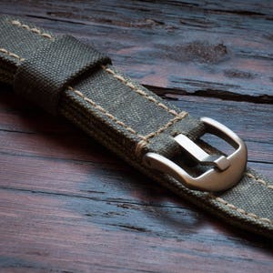 May include: Close-up of a watch strap made of olive green canvas with beige stitching. The strap features a silver-colored buckle and is displayed on a dark wooden surface. The watch band is a replacement part for a watch.