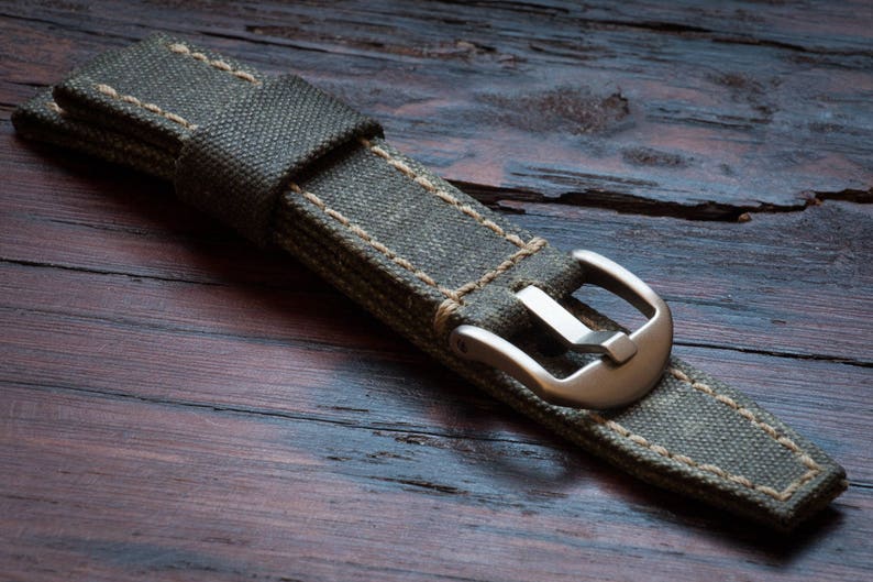 May include: Close-up of a watch strap made of olive green canvas with beige stitching. The strap features a silver-colored buckle and is displayed on a dark wooden surface. The watch band is a replacement part for a watch.