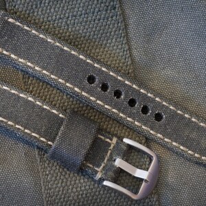 May include: A denim-style watch strap with white stitching and a silver-colored buckle. The strap has multiple holes for adjustment and is designed to fit a watch. The background is a textured, gray fabric.