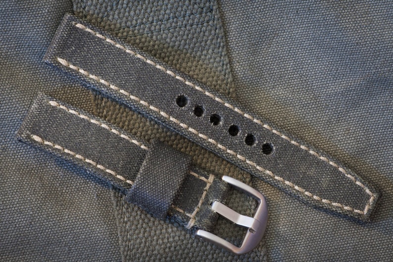 May include: A denim-style watch strap with white stitching and a silver-colored buckle. The strap has multiple holes for adjustment and is designed to fit a watch. The background is a textured, gray fabric.